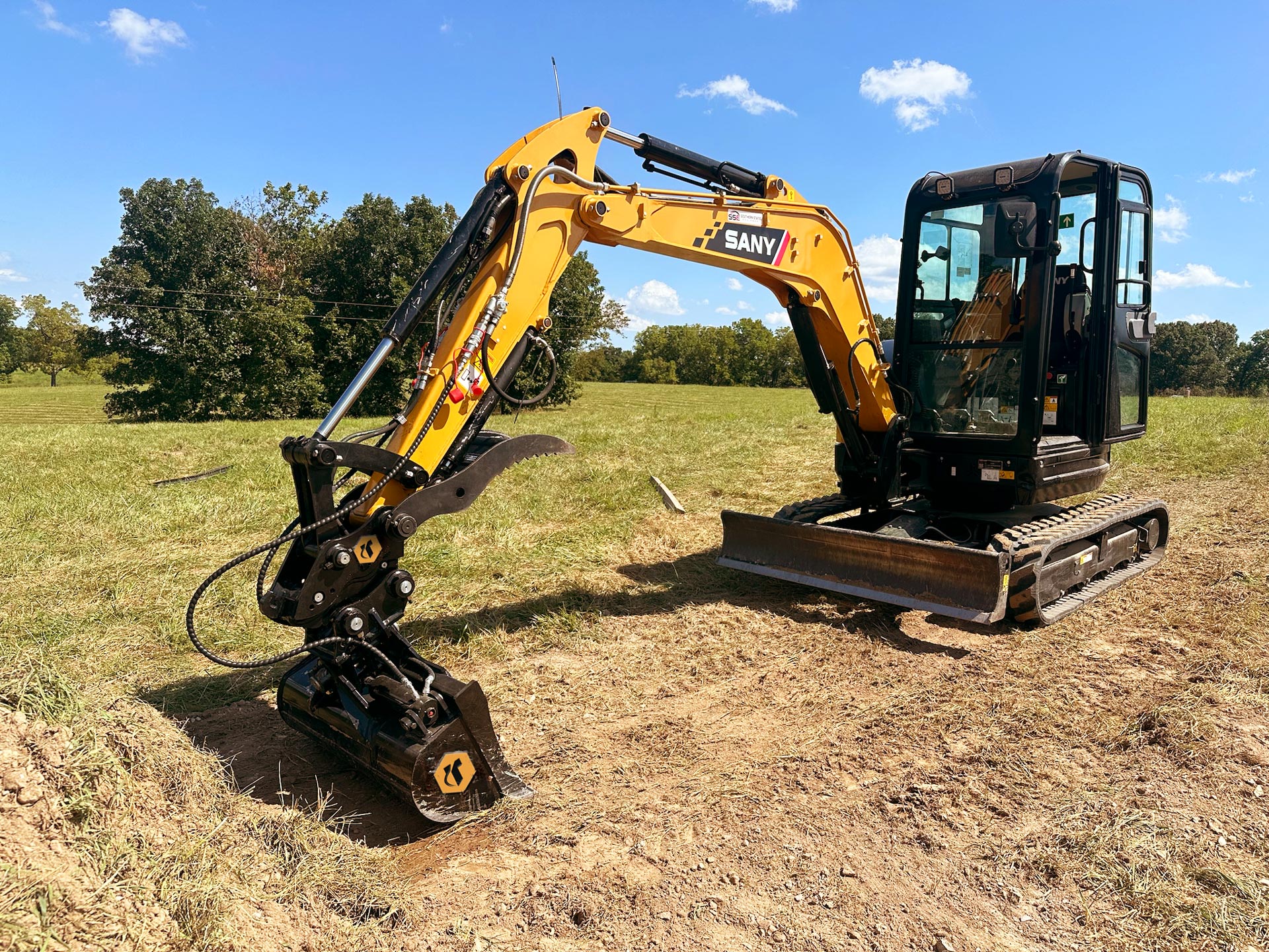 A Sany brand mini excavator digging with a tilt bucket and a thumb attached with a coupler.
